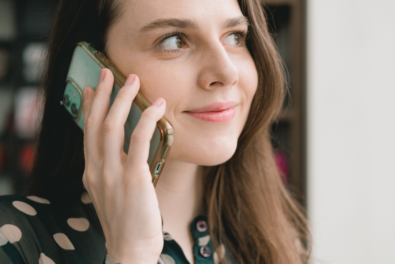 Crop young happy female millennial with long dark hair smiling and looking away while having phone conversation near window on sunny day